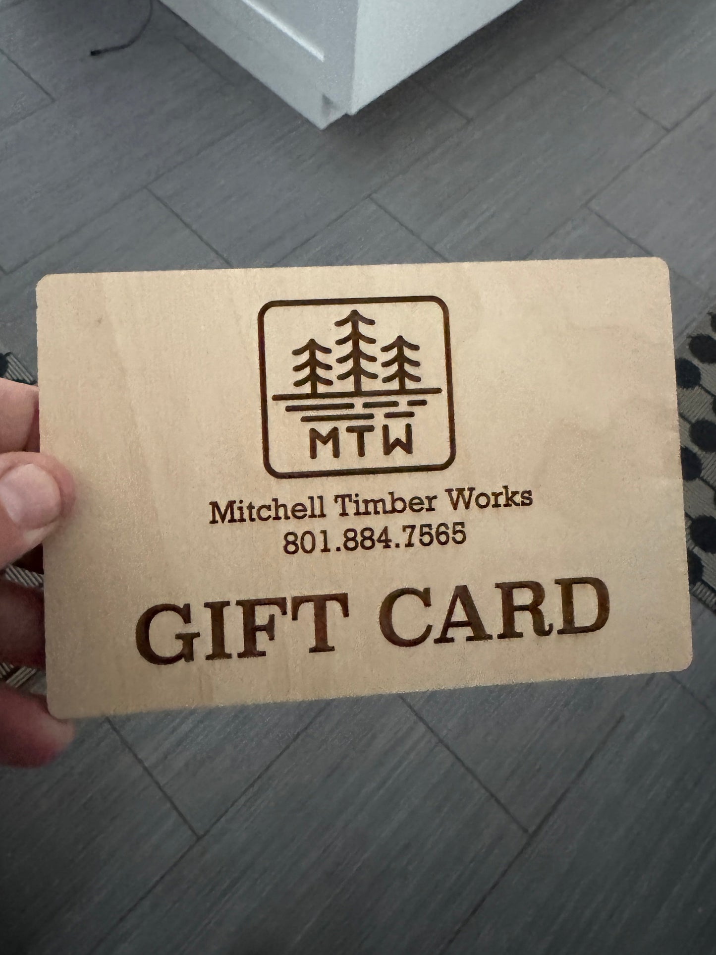 Wooden gift card with engraved text held by a hand on a tiled floor.